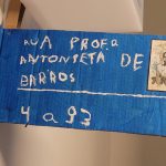 "Placa improvisada feita de papelão azul com inscrições brancas. O texto diz 'Rua Profª Antonieta de Barros' com um número '4 a 93' na parte inferior. Há uma pequena imagem de uma mulher (presumivelmente Antonieta de Barros) no canto superior direito da placa. A placa está pendurada em uma parede interior."