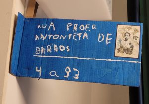 "Placa improvisada feita de papelão azul com inscrições brancas. O texto diz 'Rua Profª Antonieta de Barros' com um número '4 a 93' na parte inferior. Há uma pequena imagem de uma mulher (presumivelmente Antonieta de Barros) no canto superior direito da placa. A placa está pendurada em uma parede interior."