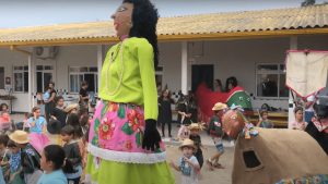 Crianças participando de uma festa ao ar livre, com música e fantasias. No centro, destaca-se uma figura de boneco gigante, uma maricota, com vestido verde e saia floral, usado como parte de uma apresentação. Ao fundo, há uma estrutura de prédio com telhado amarelo. A cena é festiva e cultural.