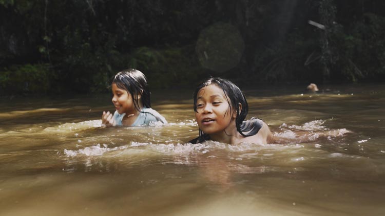 Duas crianças indígenas nadando em um rio de águas escuras, cercado por vegetação densa. Ambas estão sorrindo e se divertindo enquanto nadam. A luz do sol penetra pelas árvores, iluminando suavemente a cena.