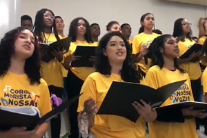 Um grupo de jovens participa de um coral, todos usando camisetas amarelas com o logotipo "Coral Marista Nossas Vozes" em azul e preto. Eles estão de pé em uma sala clara, segurando pastas pretas de partituras enquanto cantam. A expressão no rosto dos cantores reflete concentração e emoção. Há uma variedade de tonalidades de pele e tipos de cabelo entre os participantes, o que reforça a diversidade do grupo.