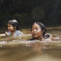 Foto de duas crianças brincando e nadando em um rio de águas marrons, em um ambiente cercado por vegetação densa e sombreada. As crianças parecem estar se divertindo e estão parcialmente submersas, com sorrisos leves no rosto.