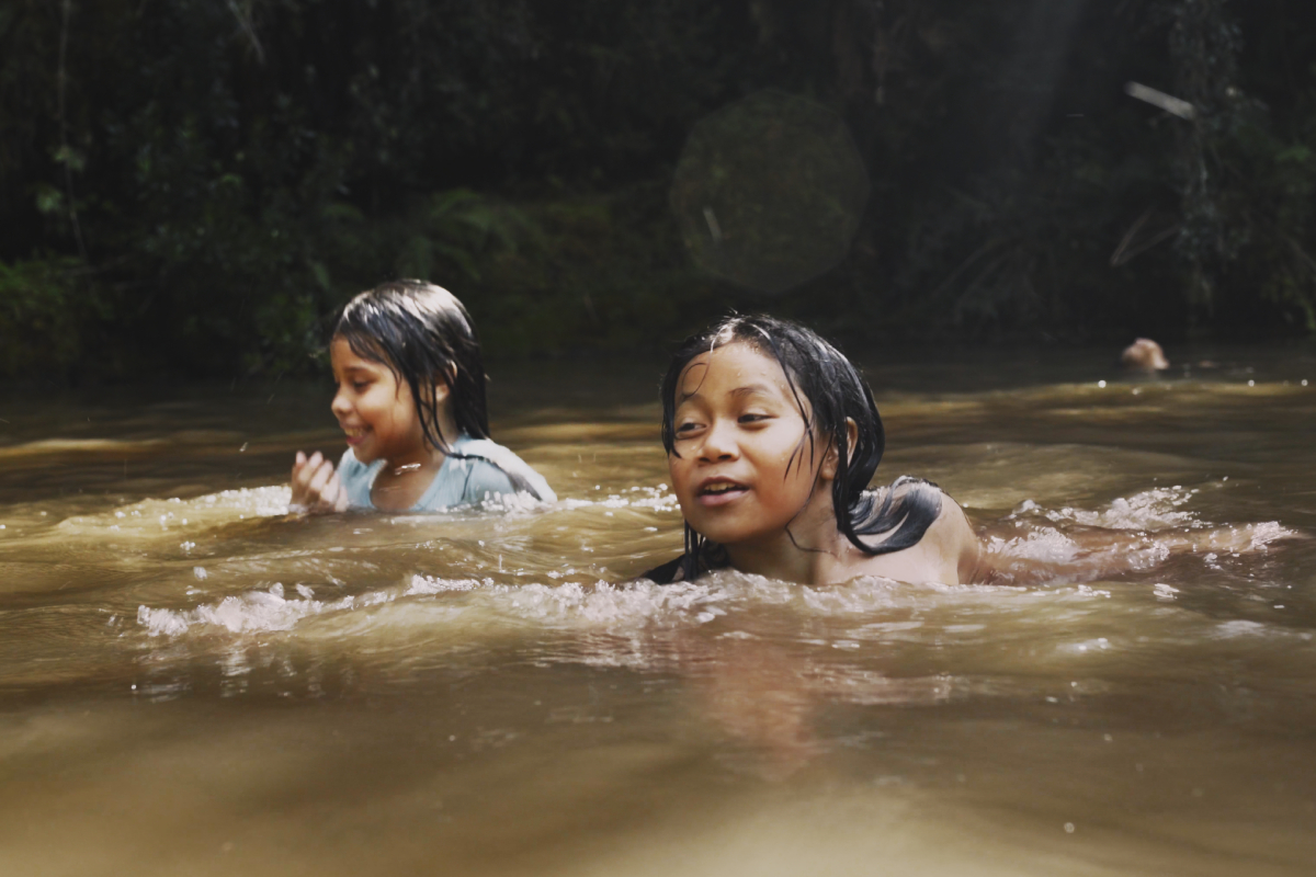 Foto de duas crianças brincando e nadando em um rio de águas marrons, em um ambiente cercado por vegetação densa e sombreada. As crianças parecem estar se divertindo e estão parcialmente submersas, com sorrisos leves no rosto.