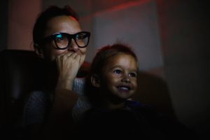 Uma mulher e uma criança sentadas em um ambiente escuro, possivelmente assistindo a um filme. Ambas sorriem e demonstram atenção ao que estão assistindo. A luz da tela ilumina parcialmente seus rostos.