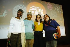 Quatro pessoas sorridentes, posicionadas lado a lado em frente a uma tela com a arte da “22ª Mostra de Cinema Infantil de Florianópolis”. As roupas e expressões sugerem um clima descontraído e amigável.