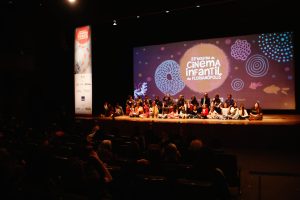 Palco de um teatro com uma projeção colorida ao fundo com o texto “22ª Mostra de Cinema Infantil de Florianópolis”. No palco, várias pessoas, principalmente crianças, estão sentadas lado a lado, olhando para a plateia, em um momento de interação após a exibição.