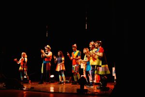 Um grupo de artistas em trajes coloridos e alegres se apresenta no palco, batendo palmas sincronizadamente. O cenário é escuro, destacando os artistas sob a luz do palco.