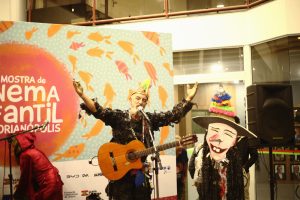 Um músico vestido com roupas coloridas, segurando um violão, se apresenta em um palco com decoração da “22ª Mostra de Cinema Infantil de Florianópolis”. Ao lado dele, uma figura com uma máscara colorida e uma fantasia vibrante.