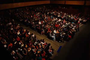 Uma grande plateia de pessoas sentadas em um teatro, observando atentamente algo no palco. A foto foi tirada de um ângulo superior, capturando a multidão em um ambiente escuro e focado.