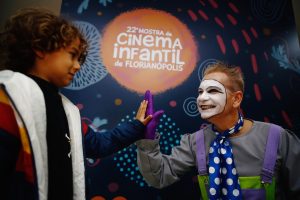 Uma criança e um artista fantasiado com maquiagem facial branca e um figurino colorido tocam as palmas das mãos em um cumprimento amigável, ambos sorrindo. Ao fundo, um banner com o logo da “22ª Mostra de Cinema Infantil de Florianópolis”