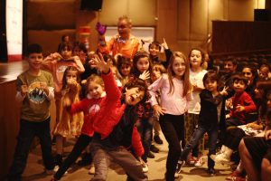 Um grupo de crianças de várias idades posa animadamente para a câmera em um teatro. Elas fazem diferentes poses expressivas e sorriem, transmitindo alegria e diversão.