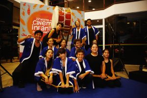 Grupo de jovens e crianças posando para uma foto em frente a um grande tambor. Eles usam trajes tradicionais japoneses azuis e brancos. Ao fundo, um banner da “22ª Mostra de Cinema Infantil de Florianópolis”.