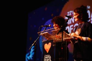 Duas jovens apresentam-se em um palco com microfones, lendo papéis em um púlpito transparente. Ao fundo, uma projeção colorida com o texto “22ª Mostra de Cinema Infantil de Florianópolis”.