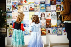 Duas meninas pequenas, usando vestidos coloridos, olham para um mural cheio de pôsteres de filmes. Elas parecem estar interessadas e admirando os cartazes.