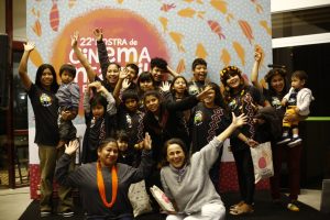 Um grupo de crianças e adultos sorridentes posando em frente a um banner da “22ª Mostra de Cinema Infantil de Florianópolis”. Alguns dos presentes usam acessórios como cocares e adereços tribais, e todos parecem animados e alegres.