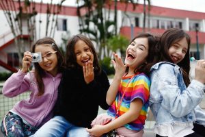 Quatro meninas sorridentes posando ao ar livre. Uma delas segura uma câmera fotográfica, enquanto as outras fazem gestos divertidos e descontraídos. Ao fundo, uma construção com detalhes em vermelho e branco.