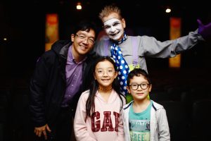 Uma família com duas crianças e um adulto posando ao lado de um artista maquiado com rosto pintado de branco, vestindo figurino colorido. Todos sorriem para a câmera, em um ambiente de teatro.