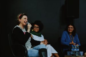 Três mulheres estão sentadas em cadeiras. A mulher à esquerda segura um microfone e fala, sorrindo. Ela tem pele clara, veste uma blusa cinza com casaco preto e calça jeans, e usa uma tiara vermelha no cabelo. Ao centro, uma mulher negra de cabelo crespo e volumoso, com óculos de armação escura, veste uma blusa branca com cachecol verde e sorri, observando a conversa. À direita, outra mulher de óculos e blusa azul observa, sorrindo. À sua frente, sobre uma mesa, há três garrafas de água e copos de plástico. O fundo é escuro.