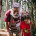 Um homem idoso de barba branca, vestindo boné e avental, está ao lado de uma criança de cabelos cacheados e camiseta vermelha. Ambos observam algo com atenção, cercados por galhos de árvore em um ambiente ao ar livre, com um bosque de árvores altas ao fundo. O homem parece concentrado e curioso, enquanto a criança olha com interesse.