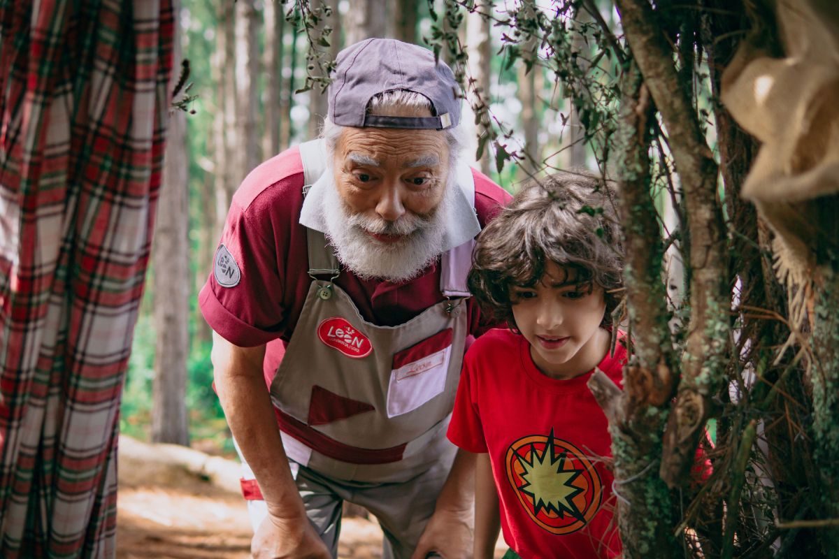 Um homem idoso de barba branca, vestindo boné e avental, está ao lado de uma criança de cabelos cacheados e camiseta vermelha. Ambos observam algo com atenção, cercados por galhos de árvore em um ambiente ao ar livre, com um bosque de árvores altas ao fundo. O homem parece concentrado e curioso, enquanto a criança olha com interesse.