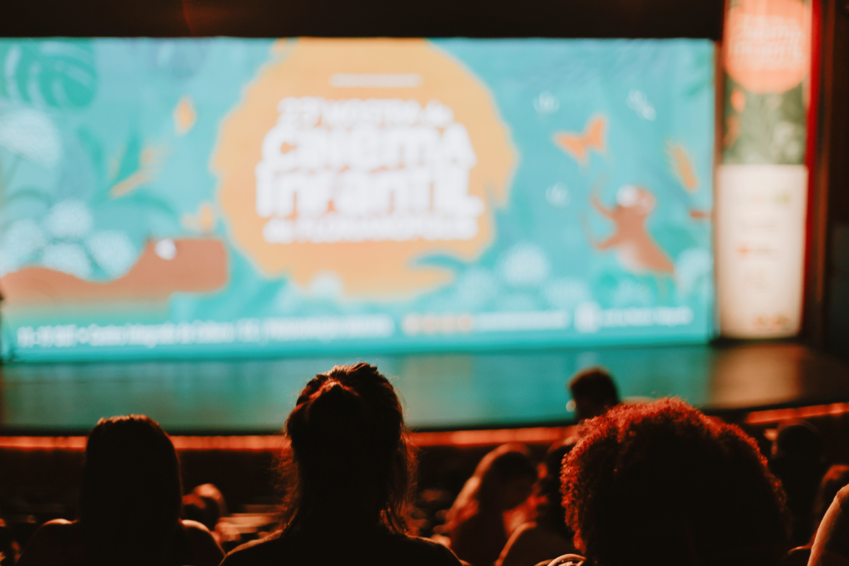 Vista de uma sala de cinema com algumas pessoas sentadas, de costas para a câmera, assistindo à projeção de uma tela que exibe o logo da "vigésima terceira Mostra de Cinema Infantil de Florianópolis". A tela de projeção é decorada com elementos infantis, como borboletas e um animal brincando, em tons de azul e laranja.