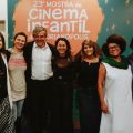 Fotografia de sete pessoas sorrindo, lado a lado, em frente a um painel que anuncia a "Vígésiam Terceira Mostra de Cinema Infantil de Florianópolis". Elas estão em pé, posando para a foto. À esquerda, uma mulher veste um blazer branco e saia listrada; ao seu lado, uma pessoa com camiseta cinza e casaco preto. O homem ao centro usa terno e camisa branca. As outras quatro mulheres à direita estão com roupas casuais, como blusas de manga longa e vestidos, uma delas com um cachecol verde sobre um vestido branco.