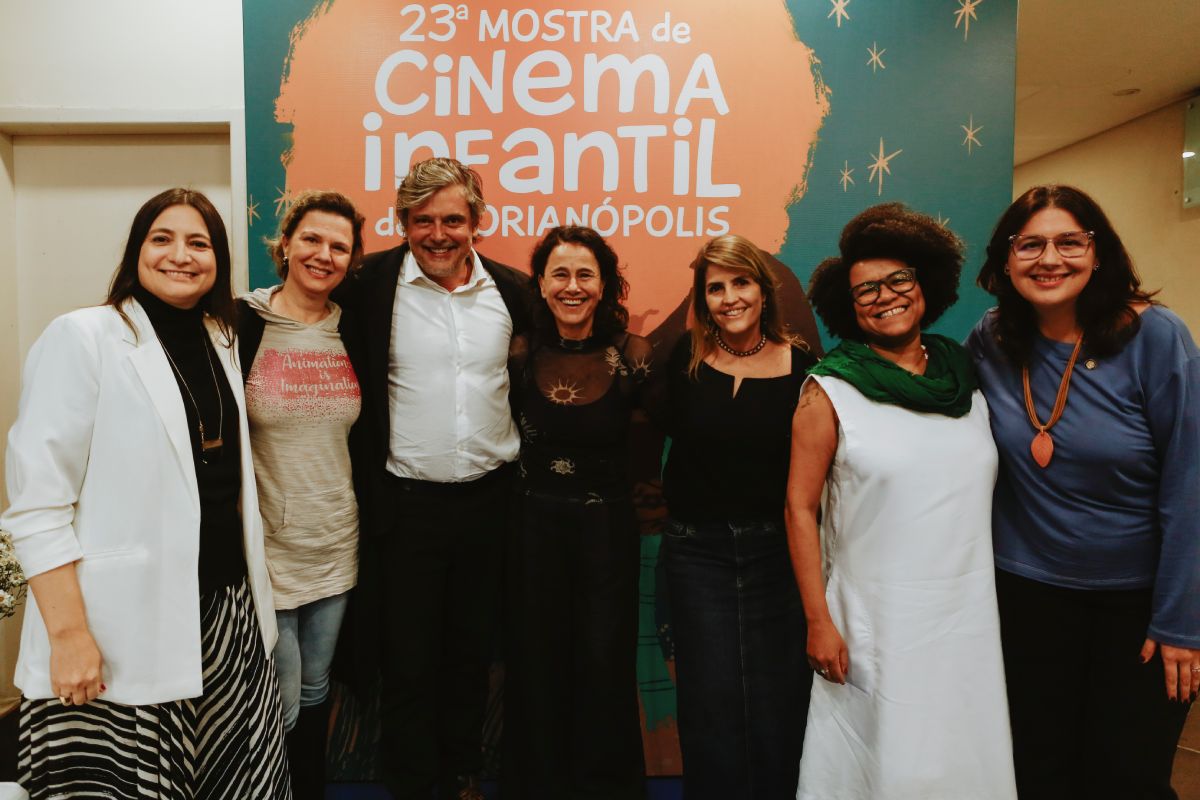 Fotografia de sete pessoas sorrindo, lado a lado, em frente a um painel que anuncia a "Vígésiam Terceira Mostra de Cinema Infantil de Florianópolis". Elas estão em pé, posando para a foto. À esquerda, uma mulher veste um blazer branco e saia listrada; ao seu lado, uma pessoa com camiseta cinza e casaco preto. O homem ao centro usa terno e camisa branca. As outras quatro mulheres à direita estão com roupas casuais, como blusas de manga longa e vestidos, uma delas com um cachecol verde sobre um vestido branco.