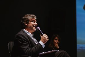 Fotografia de um homem falando ao microfone durante um evento. Ele está sentado, vestindo terno preto e camisa branca, e segura papéis na mão esquerda. Ao fundo, uma mulher desfocada, com a mão no queixo, o observa com uma expressão atenta. O fundo escuro realça a cena.