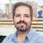 foto de Gabriel Portela: Homem sorrindo com cabelo castanho curto e barba. Ele veste uma camisa azul-claro e a foto tem um fundo com elementos urbanos desfocados.