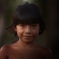 Retrato de uma criança indígena de cabelos escuros e lisos, com um leve sorriso. Ela está sem camisa e usa pequenos brincos pretos. O fundo desfocado mostra uma paisagem natural, sugerindo que a foto foi tirada ao ar livre, com iluminação suave que destaca seu rosto. A expressão da criança transmite serenidade e doçura.