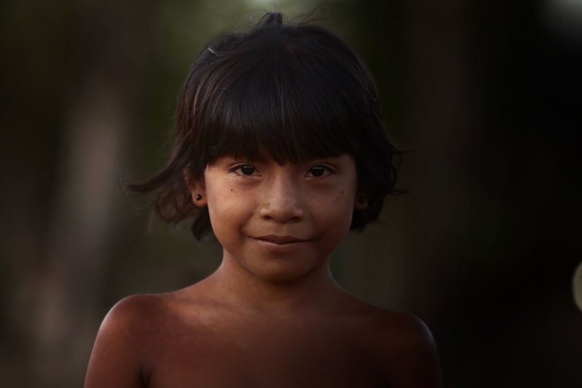 Retrato de uma criança indígena de cabelos escuros e lisos, com um leve sorriso. Ela está sem camisa e usa pequenos brincos pretos. O fundo desfocado mostra uma paisagem natural, sugerindo que a foto foi tirada ao ar livre, com iluminação suave que destaca seu rosto. A expressão da criança transmite serenidade e doçura.