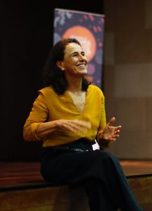 Mulher de cabelos escuros e ondulados, sorrindo, sentada em uma superfície de madeira. Ela veste uma blusa mostarda de mangas compridas e calça preta. Ao fundo, há um painel desfocado com tons de laranja e roxo. A iluminação foca na mulher, destacando sua expressão alegre.