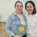 Duas mulheres estão abraçadas e sorrindo em uma sala. A mulher à esquerda usa uma jaqueta jeans sobre uma blusa listrada e segura um material gráfico, que é um cartaz da "23ª Mostra de Cinema Infantil de Florianópolis". A mulher à direita veste uma blusa branca e tem cabelo ondulado e solto. O ambiente ao fundo tem cadeiras azuis, piso de madeira e uma parede clara, sugerindo um espaço de eventos ou reunião. A expressão das duas mulheres transmite alegria e camaradagem.