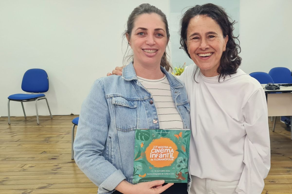 Duas mulheres estão abraçadas e sorrindo em uma sala. A mulher à esquerda usa uma jaqueta jeans sobre uma blusa listrada e segura um material gráfico, que é um cartaz da "23ª Mostra de Cinema Infantil de Florianópolis". A mulher à direita veste uma blusa branca e tem cabelo ondulado e solto. O ambiente ao fundo tem cadeiras azuis, piso de madeira e uma parede clara, sugerindo um espaço de eventos ou reunião. A expressão das duas mulheres transmite alegria e camaradagem.