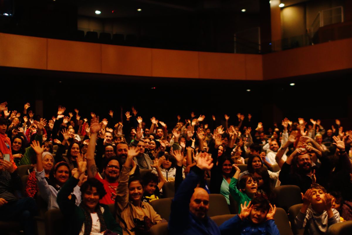 Auditório cheio de pessoas, todas sorrindo e levantando as mãos. O ambiente está escuro, com iluminação suave destacando o público, que parece participar de uma interação coletiva. A maioria das pessoas aparenta estar animada, com diferentes idades e expressões de entusiasmo.