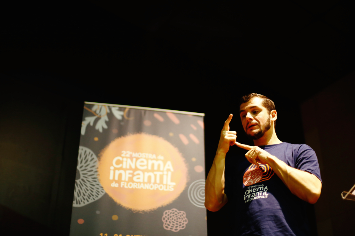 Fotografia de um homem utilizando Língua Brasileira de Sinais (Libras) durante um evento. Ele veste uma camiseta azul com o logo da "Vigésima segunda Mostra de Cinema Infantil de Florianópolis". Ao fundo, há um banner com o mesmo logo, destacando a frase "Vigésima segunda Mostra de Cinema Infantil de Florianópolis", e um design colorido em tons de laranja, branco e cinza. O ambiente parece ser interno, com iluminação focalizada.