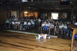 Crianças sentadas em um auditório assistindo a uma exibição de filme projetado em uma tela. A projeção mostra uma animação colorida. Alguns bonecos de personagens estão posicionados na frente do palco, à vista das crianças. O auditório é o mesmo das imagens anteriores, com uma bandeira da Unibave pendurada na parede.