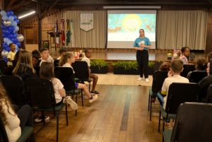 Crianças estão sentadas em cadeiras no auditório, algumas olhando para trás em direção à câmera, enquanto outras observam uma mulher de pé no palco. Ela usa uma camiseta azul da "Mostra de Cinema Infantil de Florianópolis" e parece se comunicar com as crianças. No palco, há bonecos de personagens infantis, e atrás dela está a tela de projeção com o logo do evento.