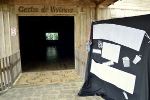 Entrada do "Centro de Vivência", com uma placa de madeira acima da porta. Ao lado da entrada, há um painel decorado com recortes temáticos de cinema, incluindo baldes de pipoca e rolos de filme. No interior escuro, uma tela de projeção exibe uma cena de filme.