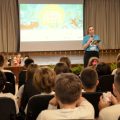 Fotografia de um auditório com várias crianças sentadas, voltadas para uma mulher em pé no palco. Ela segura um microfone e um tablet, vestindo uma camiseta azul com o logo da "Mostra de Cinema Infantil de Florianópolis". Ao fundo, há uma tela projetada com a arte colorida do evento. Sobre o palco, estão posicionados alguns bonecos de personagens. No canto superior esquerdo, vê-se um banner da universidade Unibave.