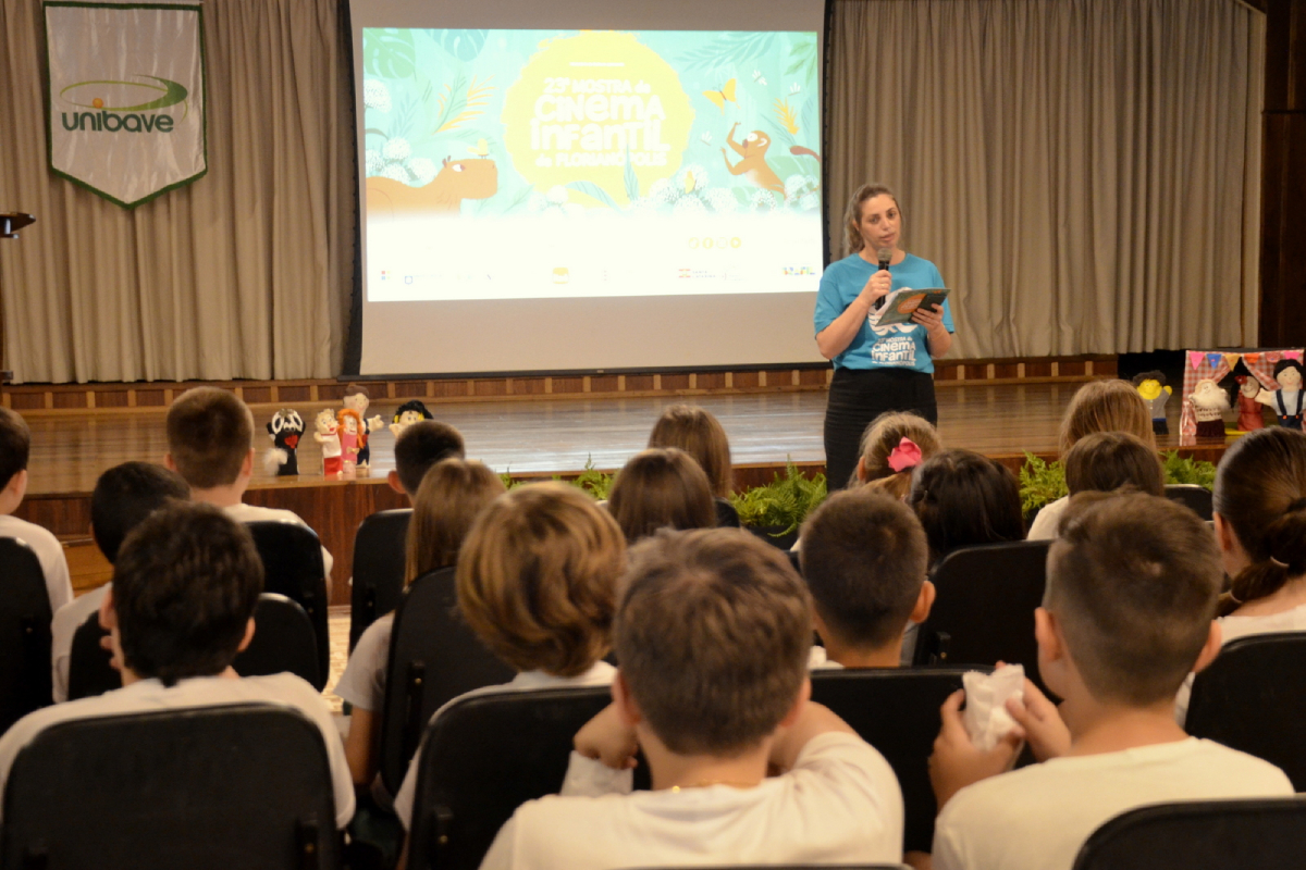 Fotografia de um auditório com várias crianças sentadas, voltadas para uma mulher em pé no palco. Ela segura um microfone e um tablet, vestindo uma camiseta azul com o logo da "Mostra de Cinema Infantil de Florianópolis". Ao fundo, há uma tela projetada com a arte colorida do evento. Sobre o palco, estão posicionados alguns bonecos de personagens. No canto superior esquerdo, vê-se um banner da universidade Unibave.