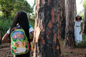 Uma fotografia de duas crianças em um ambiente arborizado. Em primeiro plano, uma criança de cabelos longos, de costas para a câmera, está usando uma mochila colorida com desenhos. Ao fundo, outra criança, vestida com um vestido branco, observa parcialmente escondida atrás de uma árvore. O cenário ao redor é composto por árvores de tronco largo e vegetação natural, transmitindo uma atmosfera de brincadeira ao ar livre.