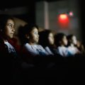 A foto mostra um grupo de crianças sentadas lado a lado em uma sala escura, como em um cinema ou teatro. No primeiro plano, uma menina com casaco vermelho e expressão atenta olha fixamente para frente, onde provavelmente está uma tela de projeção. Ao seu lado, outras crianças, parcialmente desfocadas, também assistem com concentração. A luz da tela ilumina suavemente seus rostos, destacando a curiosidade e o interesse nas expressões. Ao fundo, é possível ver um sinal de saída ("EXIT") iluminado em vermelho, contribuindo para o ambiente escuro da sala.